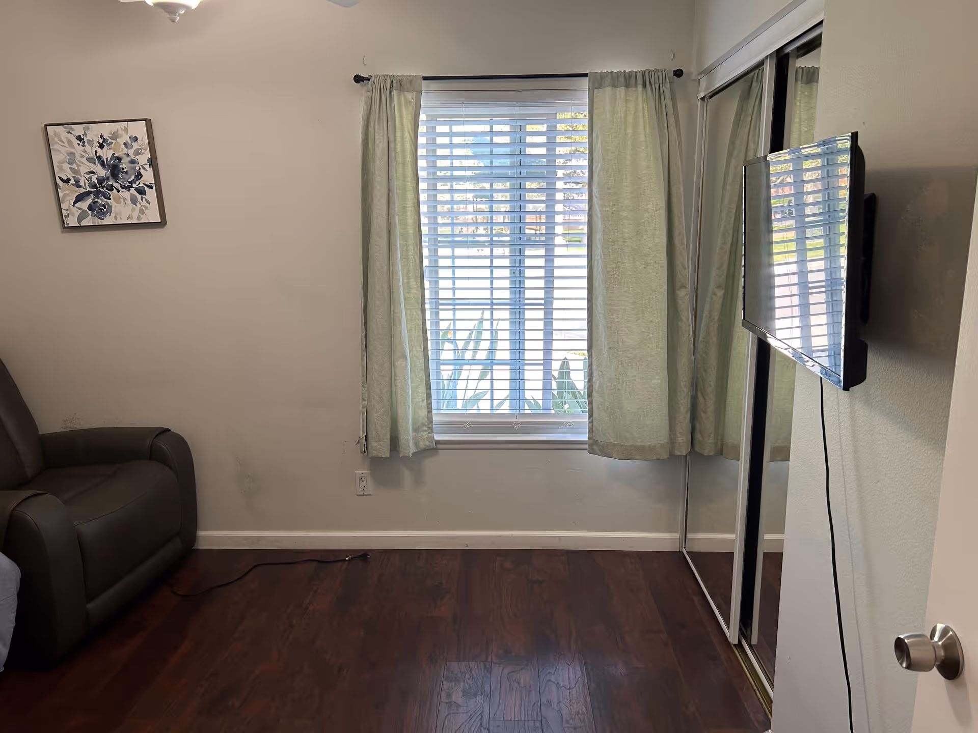 A small room with a window covered by light green curtains and white blinds. There is a brown armchair on the left side, a framed floral artwork on the wall above it, and a flat-screen TV mounted on the right wall. The floor is dark wood, and there is a mirrored sliding closet door next to the TV.