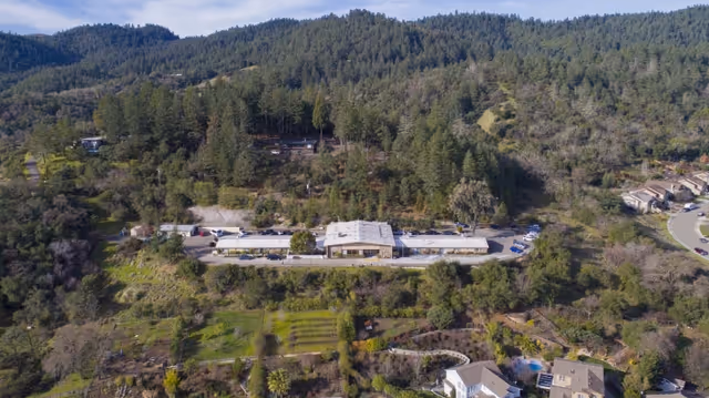 Aerial view of a long single-story healthcare building set along a hillside surrounded by trees and nearby homes.