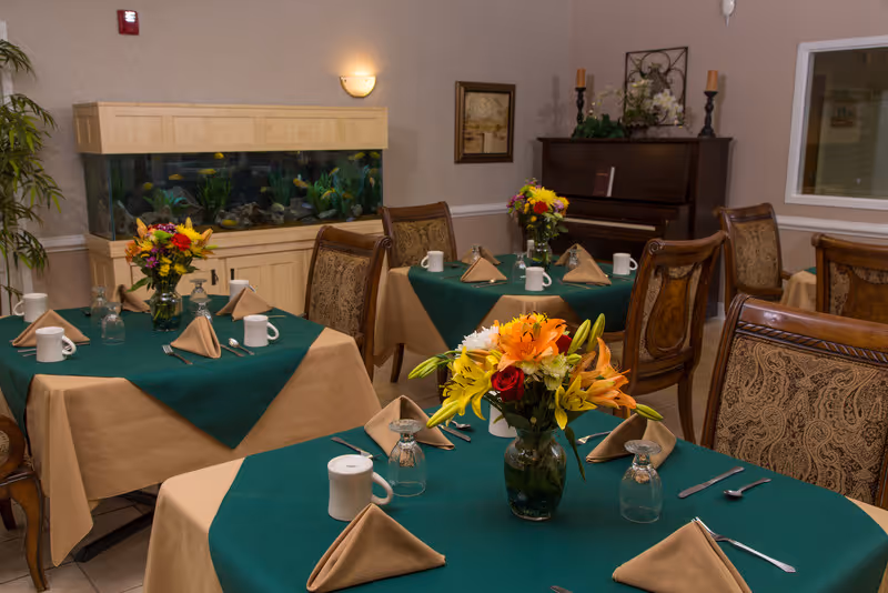 A dining room with several tables set with green tablecloths, napkins, cups and floral centerpieces, an aquarium and a piano in the background.