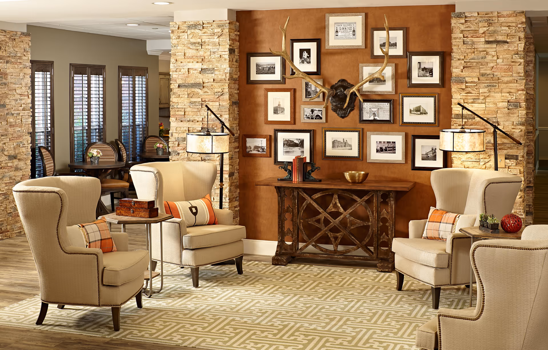 A cozy senior living common area with four beige wingback armchairs arranged around small side tables. The room features a patterned rug, stone accent walls, and a wooden console table against a brown wall decorated with framed black and white photographs and a mounted set of antlers. In the background, there are windows with wooden blinds and a dining area with tables and chairs.