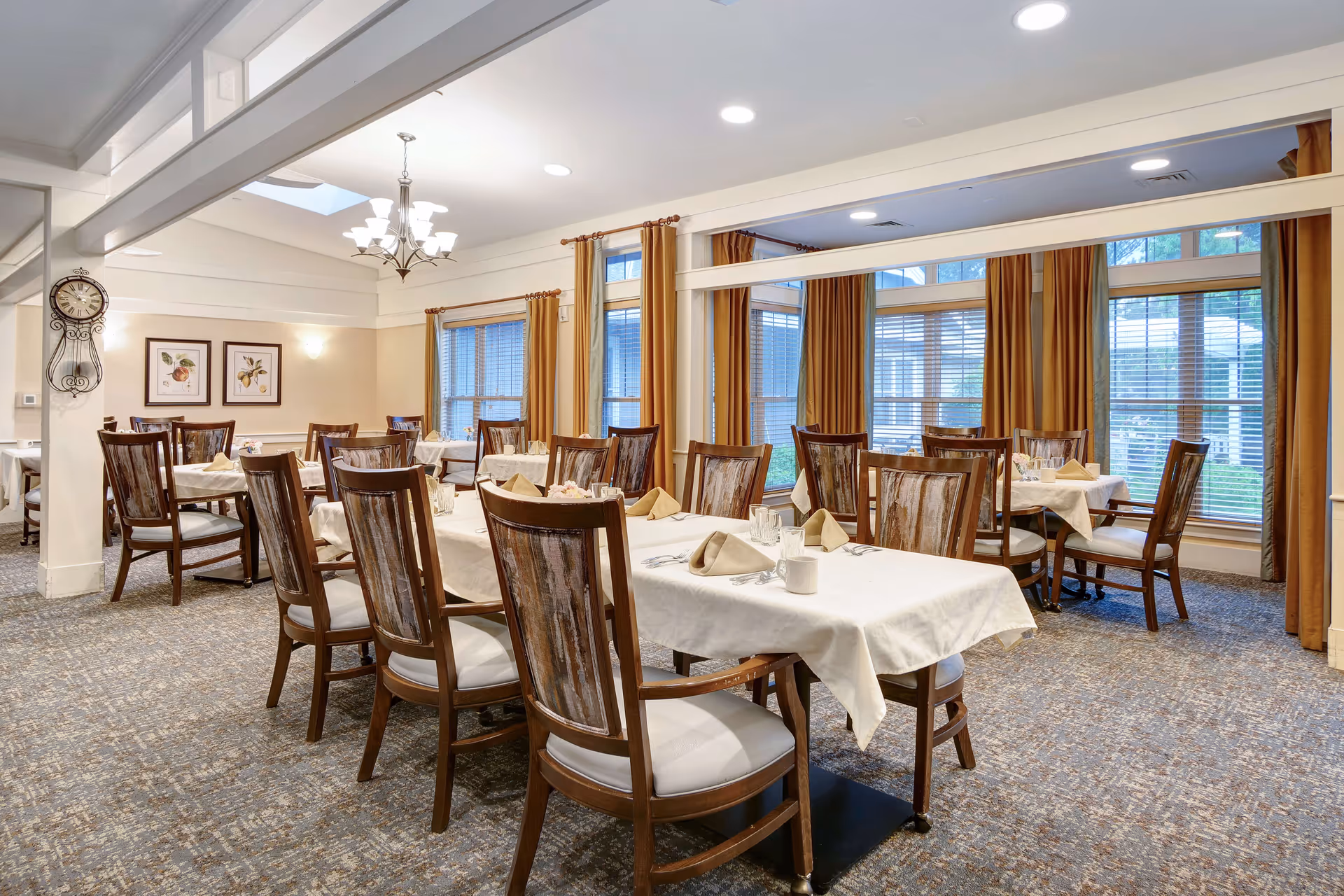 A bright dining room with several wooden tables and chairs arranged neatly. Each table is covered with a white tablecloth and set with napkins, glasses, and cups. Large windows with brown curtains allow natural light to fill the room. The walls are decorated with framed botanical prints and a large clock is mounted on one wall. The carpet has a patterned design and the ceiling features recessed lighting and a chandelier.