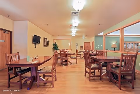 A dining room with several wooden tables and chairs arranged neatly on a wooden floor. The walls are painted in light colors with some green accents, and there is a small wall-mounted TV and framed artwork on one wall. Ceiling lights illuminate the room evenly.