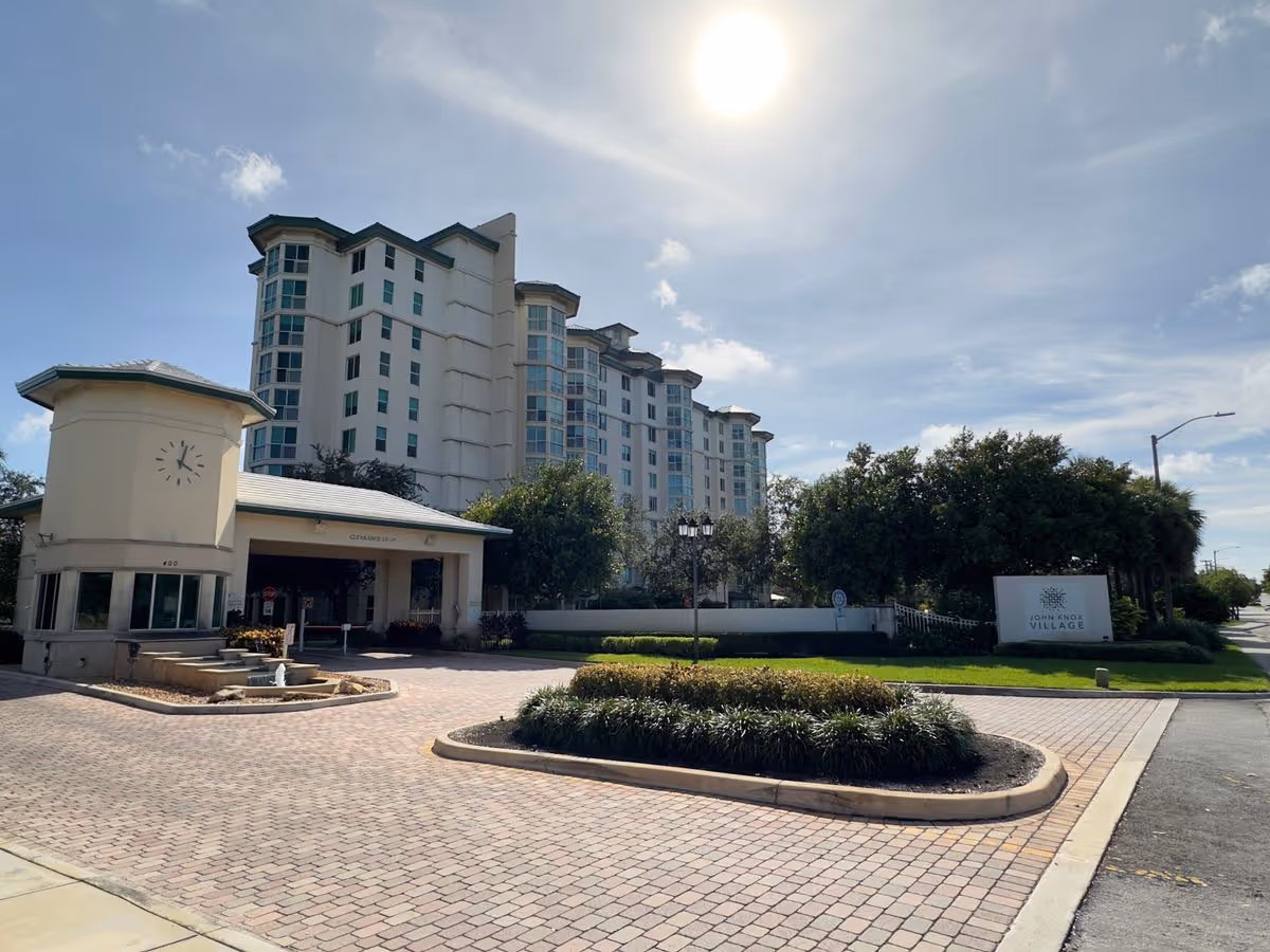 Exterior front entrance and multi-story building of a senior living campus with a porte-cochere, clock tower, landscaped roundabout, and a 'John Knox Village' sign.