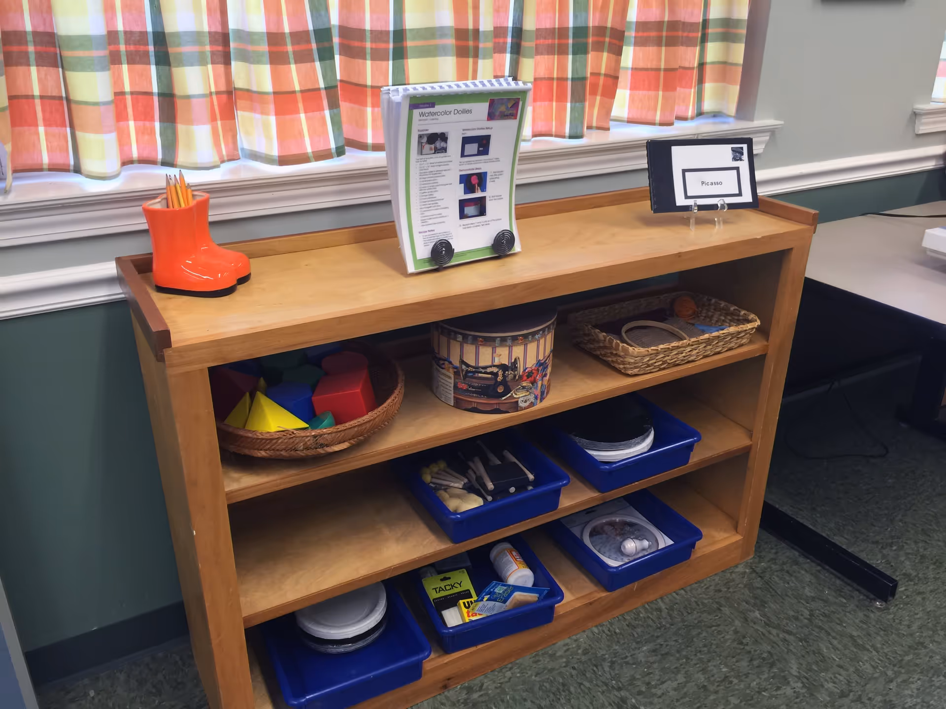 A wooden shelving unit with various craft supplies including colored pencils in an orange boot-shaped holder, geometric shapes in a basket, a round tin, and blue trays containing different art materials. The shelf is placed against a wall with green paint and white trim, beneath a window with plaid curtains in orange, green, and white colors.
