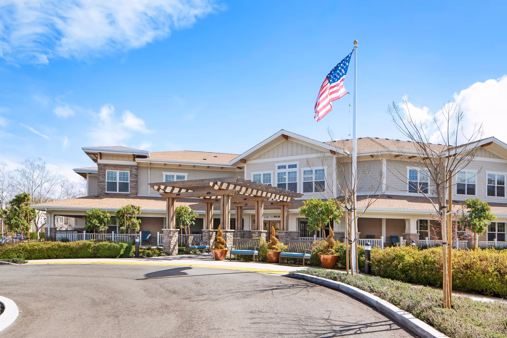 Exterior view of a two-story senior living facility building with a covered entrance featuring wooden pergola, benches, potted plants, and an American flag on a flagpole in front. The sky is clear with some clouds.