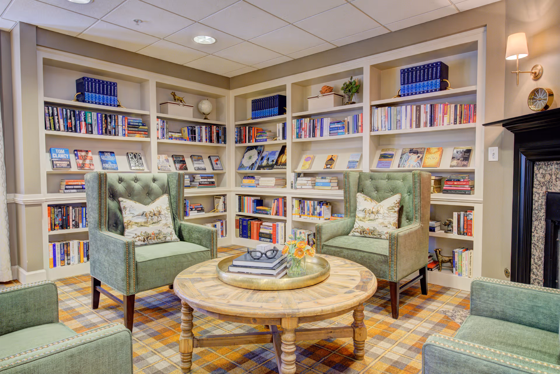 A cozy reading room with built-in bookshelves filled with books, two green upholstered armchairs with decorative pillows, a round wooden coffee table with books and a small flower arrangement, and a fireplace with a black mantel on the right side.