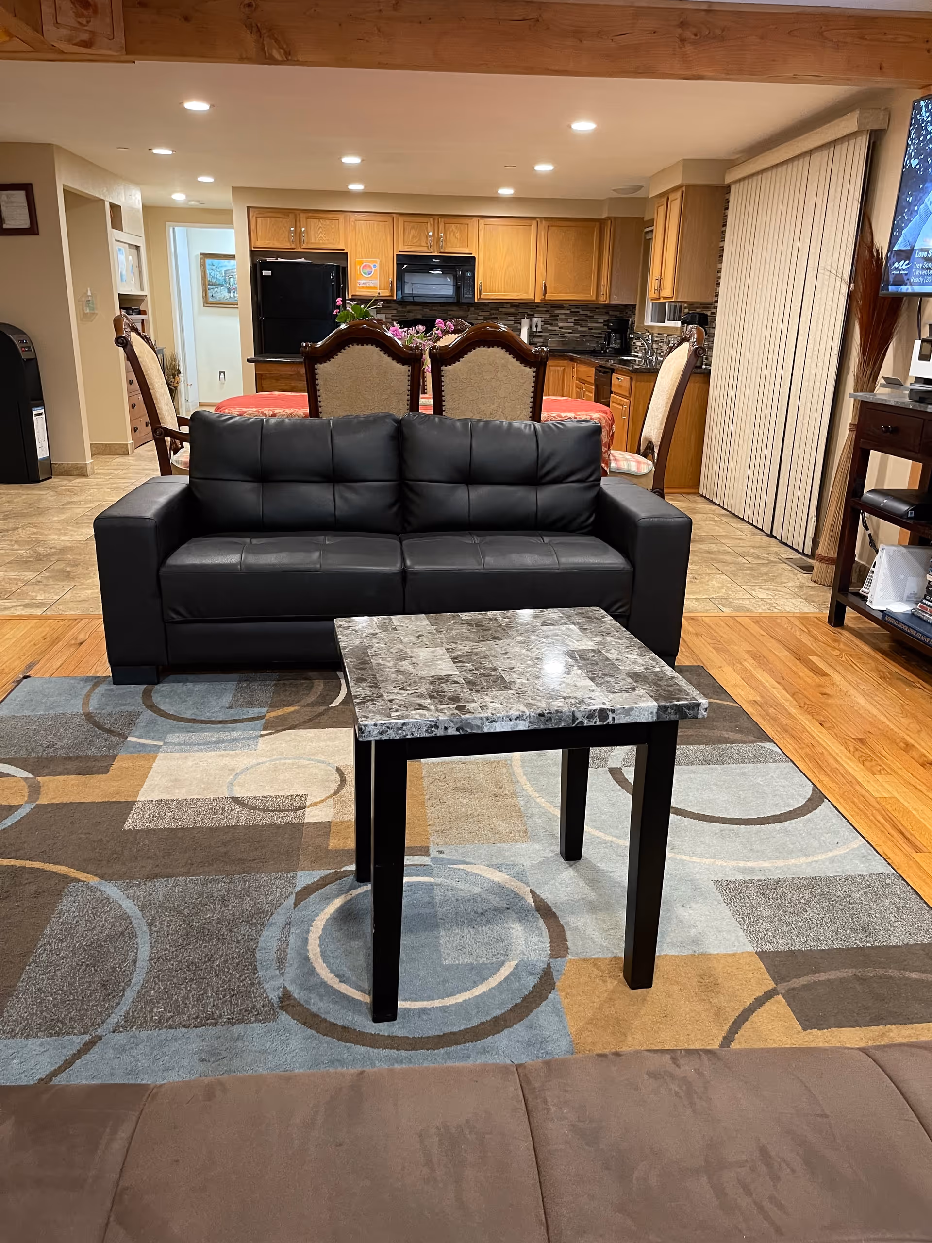 Interior view of a living area in a senior care facility showing a black leather sofa, a small square marble-top table, and a patterned area rug. Behind the sofa is a dining table with four upholstered chairs and a kitchen with wooden cabinets, a black refrigerator, and a microwave. The room has recessed lighting and a sliding door with vertical blinds.