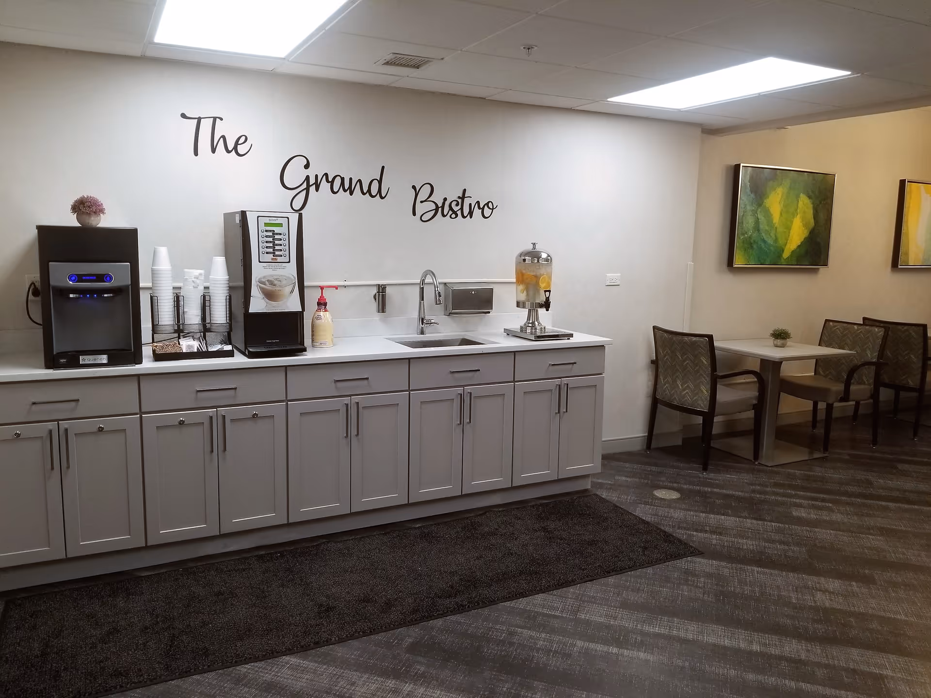 Interior view of The Grand Bistro area in a supportive living facility featuring a countertop with a coffee machine, cups, a beverage dispenser, and a sink. To the right, there are tables with chairs and colorful abstract paintings on the wall.