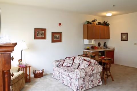A cozy living room area with a floral patterned sofa, a wooden side table with a lamp, and a small dining table with two chairs. The room has beige carpeting and white walls decorated with framed pictures. There is a kitchenette in the background with wooden cabinets, a microwave, and a small countertop with decorative items.