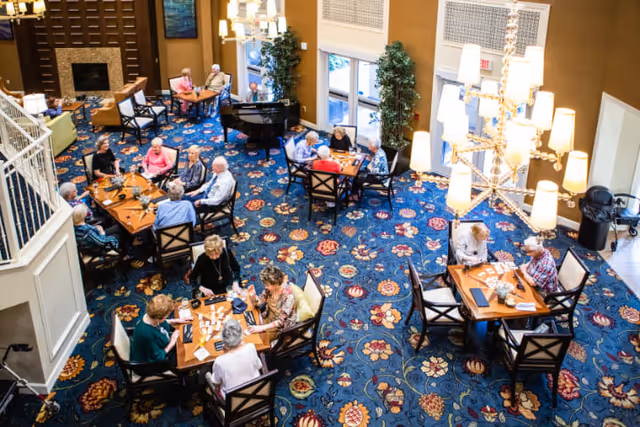 A spacious common area in a senior living facility with multiple tables where elderly residents are seated playing games and socializing. The room features a vibrant blue floral carpet, large windows letting in natural light, a grand piano, and modern chandeliers hanging from the ceiling.