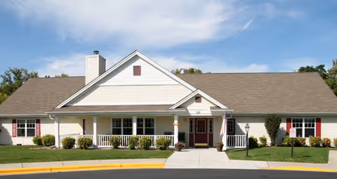 Single-story building with beige siding and a brown shingled roof under a blue sky. The building has a covered front porch with white railings, two red doors, and several windows with red shutters. There are small bushes and a well-maintained lawn in front, along with a sidewalk and a yellow curb.