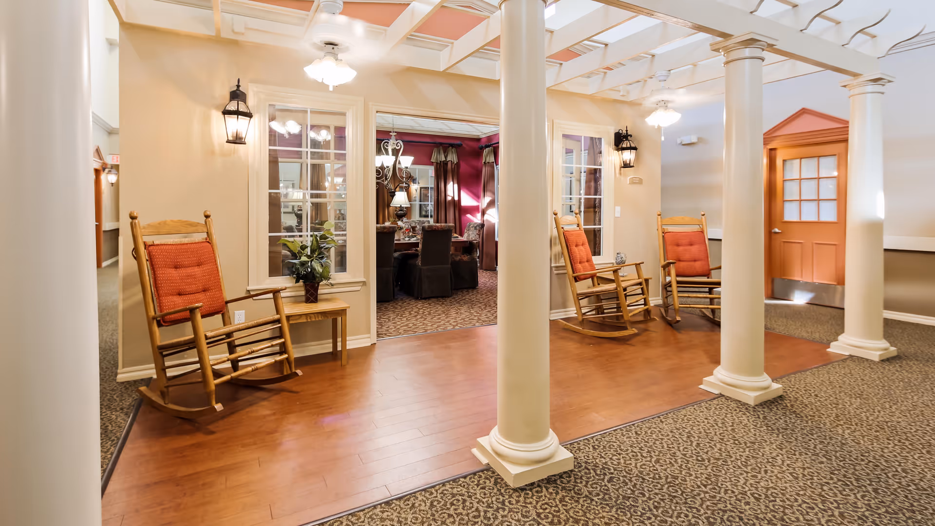 Interior view of a senior living facility hallway with four white columns and three wooden rocking chairs with red cushions placed on a wooden floor area. There are wall-mounted lantern-style lights and a small table with a plant between two chairs. In the background, a room with carpeted floor, dining chairs, and a chandelier is visible through large windows and an open doorway. A coral-colored door with glass panes is on the right side.