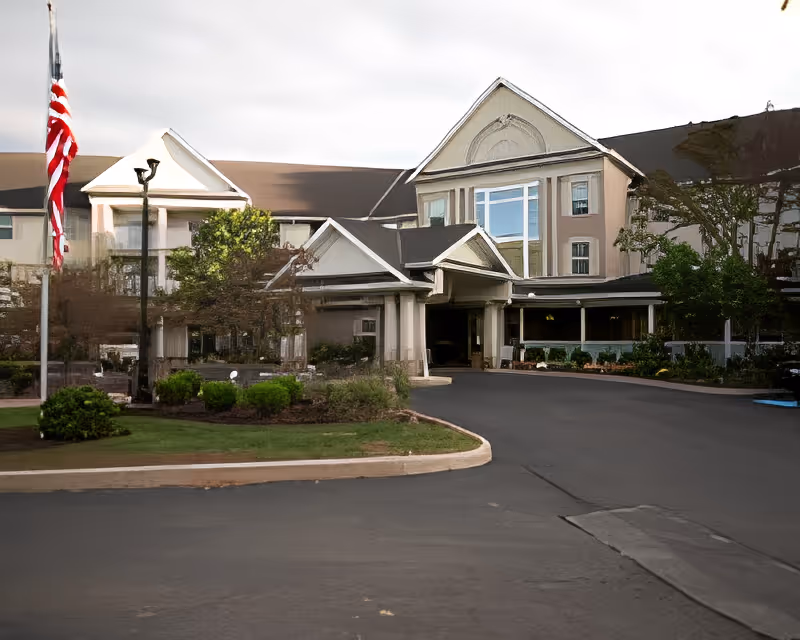 Front entrance of a multi-story senior living building with a covered porte-cochere, circular driveway, landscaped beds, and an American flag.