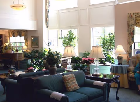 A bright and spacious living room area with large windows letting in natural light. The room features green upholstered sofas with plaid pillows, several table lamps with white shades, a grand piano in the background, and various potted plants and floral arrangements. There is also a side table with a lamp and a person partially visible sitting on a chair to the right.