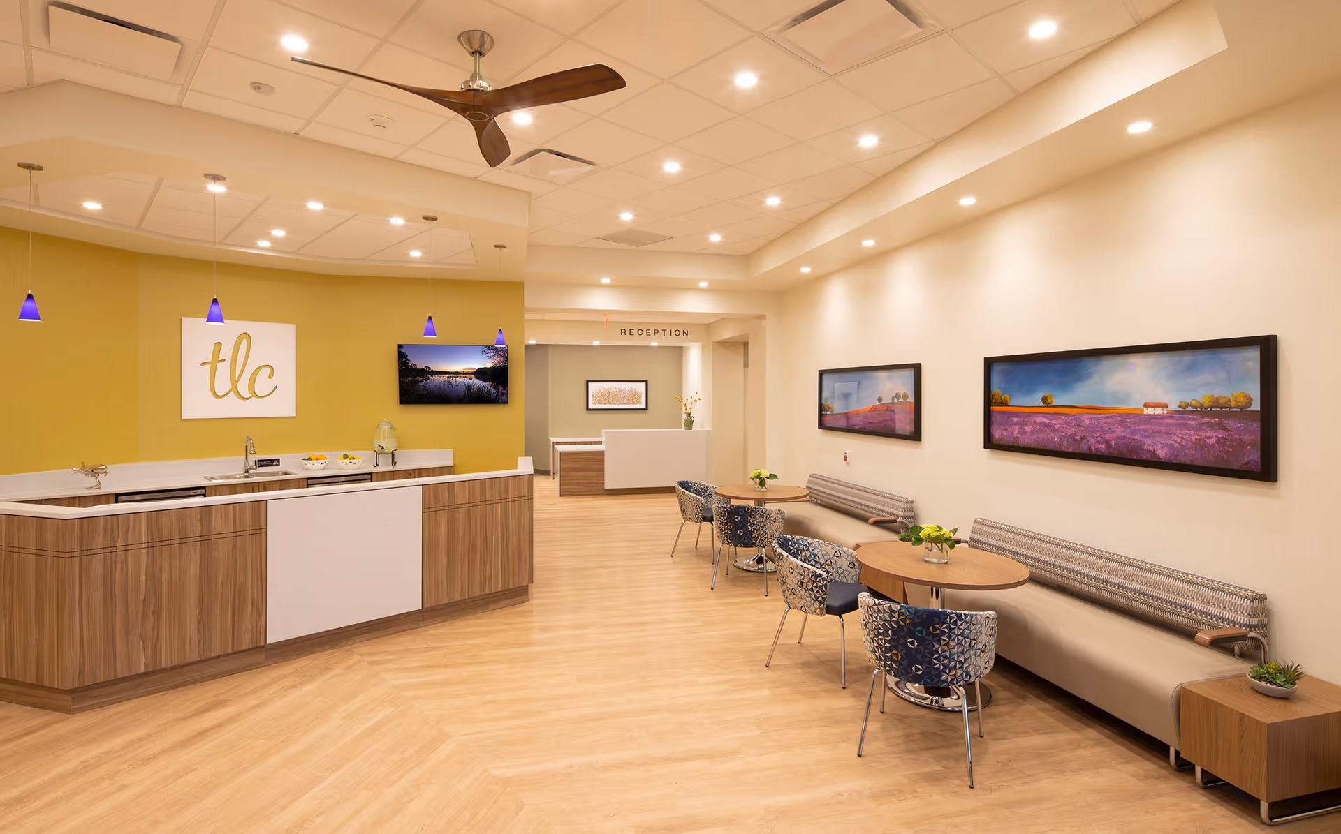 A bright and modern reception area in TLC Living Community featuring a wooden reception desk with a white countertop, three blue pendant lights hanging above, and a ceiling fan. The walls are painted yellow and cream, adorned with two landscape paintings and a TV screen. There are round tables with patterned chairs and bench seating along the wall, decorated with small plants.