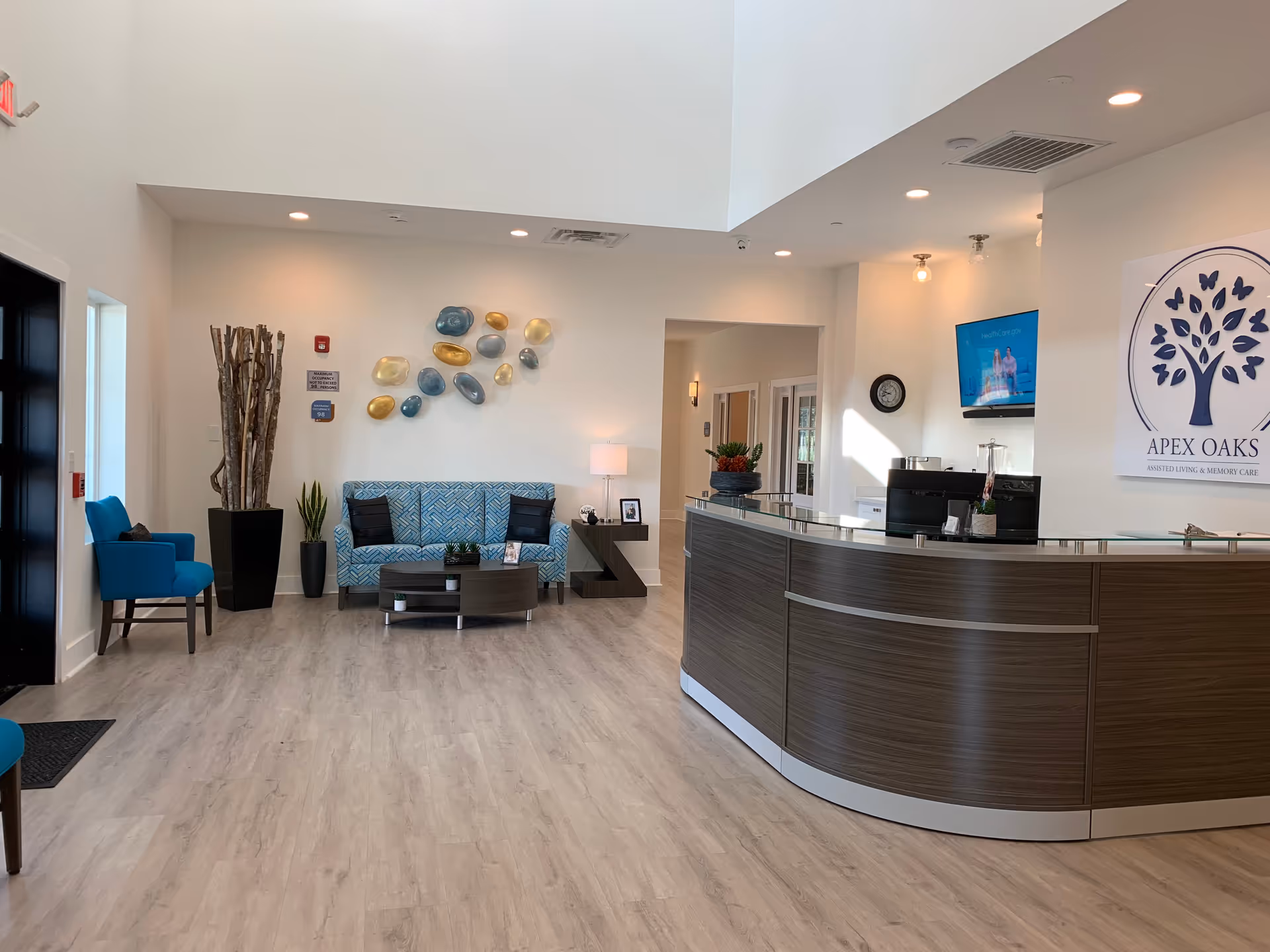 Bright, modern reception lobby with a curved front desk, seating area with blue sofa and chairs, and an Apex Oaks logo on the wall.