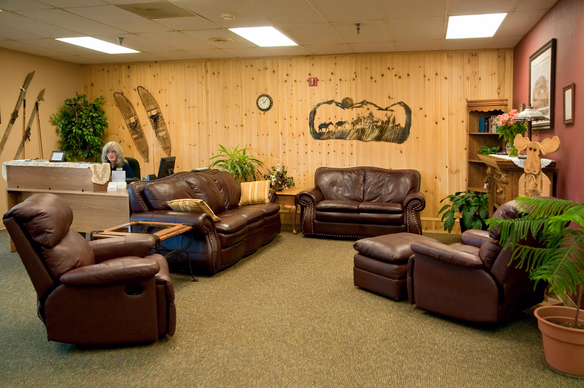 A cozy waiting or lounge area with brown leather sofas and recliners arranged around a small wooden table. The room has wood-paneled walls with decorative items including vintage snowshoes, a clock, and a metal wall art piece. There is a wooden desk with a woman seated behind it, some potted plants, a wooden bookshelf, and a wooden moose sculpture. The carpet is greenish and the ceiling has fluorescent lights.