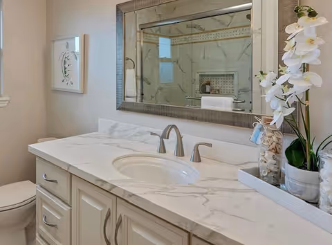 A clean and modern bathroom featuring a white marble countertop with an oval sink and silver faucet. Above the sink is a large rectangular mirror with a textured frame. To the right of the sink, there is a decorative arrangement including white orchids and a glass jar filled with seashells. In the background, a shower area with marble tiles and a towel hanging on a bar is visible. A toilet is partially visible on the left side, and a framed picture hangs on the wall above it.