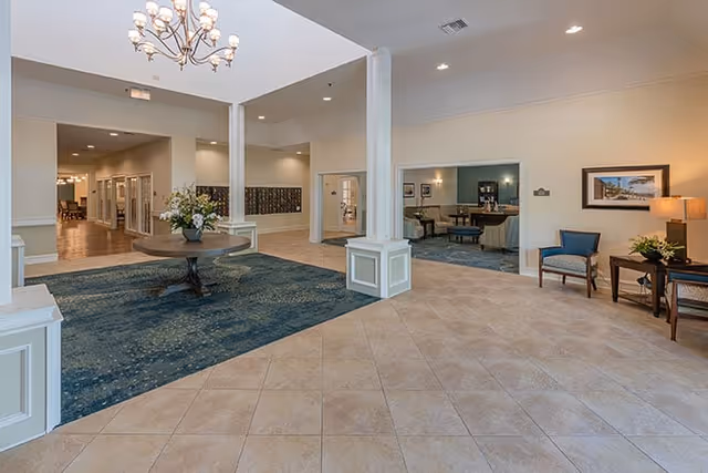Spacious and well-lit interior common area of a senior living facility with tiled floors and a large blue carpet. A round wooden table with a flower arrangement is centered on the carpet. The room features white columns, beige walls, and recessed lighting. To the right, there is a small seating area with two chairs, a side table with a lamp, and framed artwork on the wall. In the background, there are mailboxes and additional rooms visible.