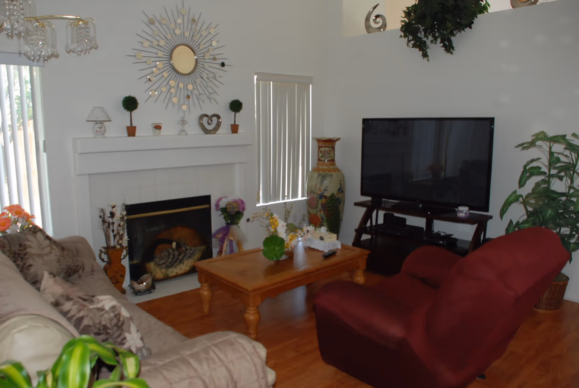 A cozy living room with a beige sofa and a red armchair facing a wooden coffee table. The room features a fireplace with decorative items on the mantel, a large flat-screen TV on a stand, and various plants and floral arrangements. A decorative sunburst mirror hangs above the fireplace, and vertical blinds cover the windows.
