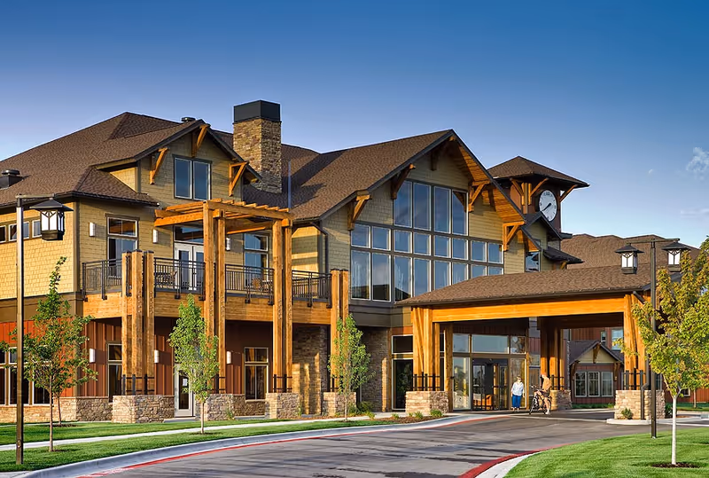 Exterior view of a large senior living facility building with wooden architectural features, large windows, a covered entrance, and a clock tower. There are a few small trees and a paved driveway leading to the entrance under a clear blue sky.