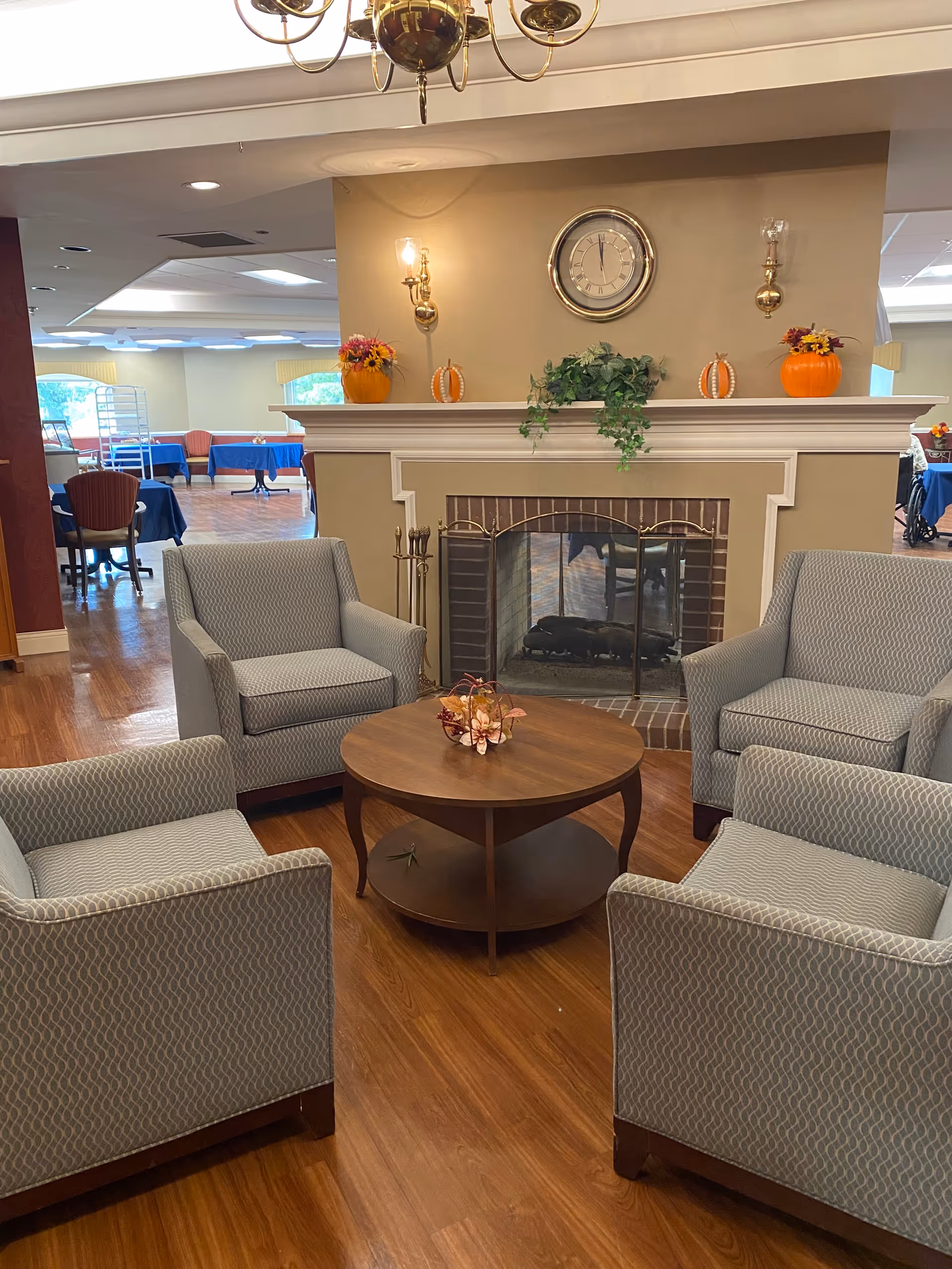 A cozy seating area with four gray patterned armchairs arranged around a round wooden coffee table with a small floral centerpiece. Behind the seating area is a fireplace with a brick interior and a white mantel decorated with two orange pumpkin decorations, two small flower arrangements, and green foliage. Above the mantel is a round clock and two wall sconces with lit bulbs. In the background, there are tables covered with blue tablecloths and chairs, suggesting a dining area.