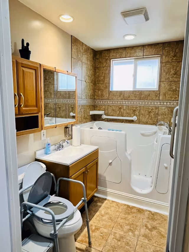 A bathroom with beige tiled walls and floor, featuring a walk-in bathtub with safety grab bars, a white toilet with a raised seat and support frame, a wooden vanity with a white sink, a wall-mounted mirror cabinet, and a small window above the bathtub.