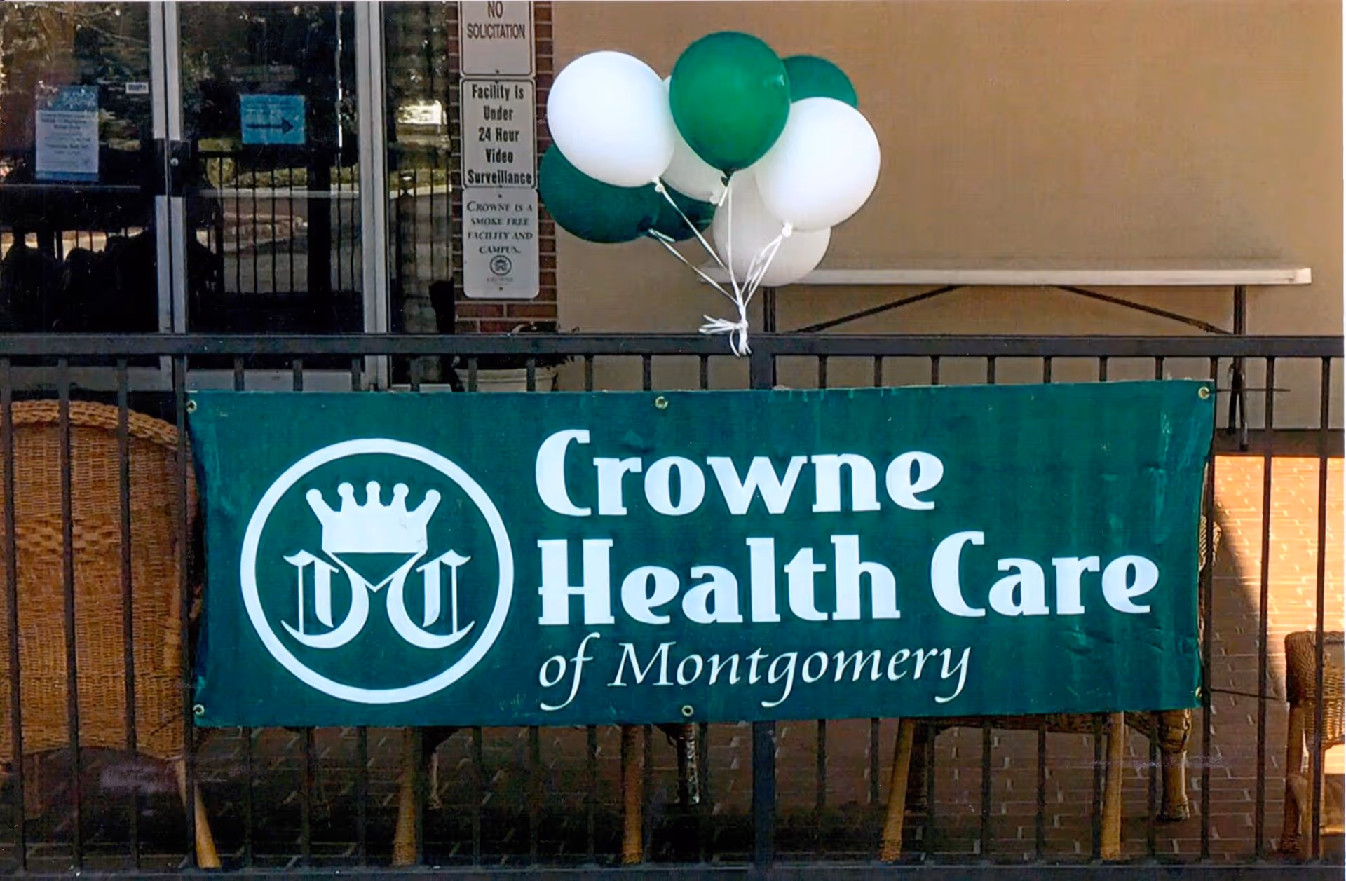 A green banner reading "Crowne Health Care of Montgomery" hangs on a railing with green and white balloons tied above it at the facility entrance.