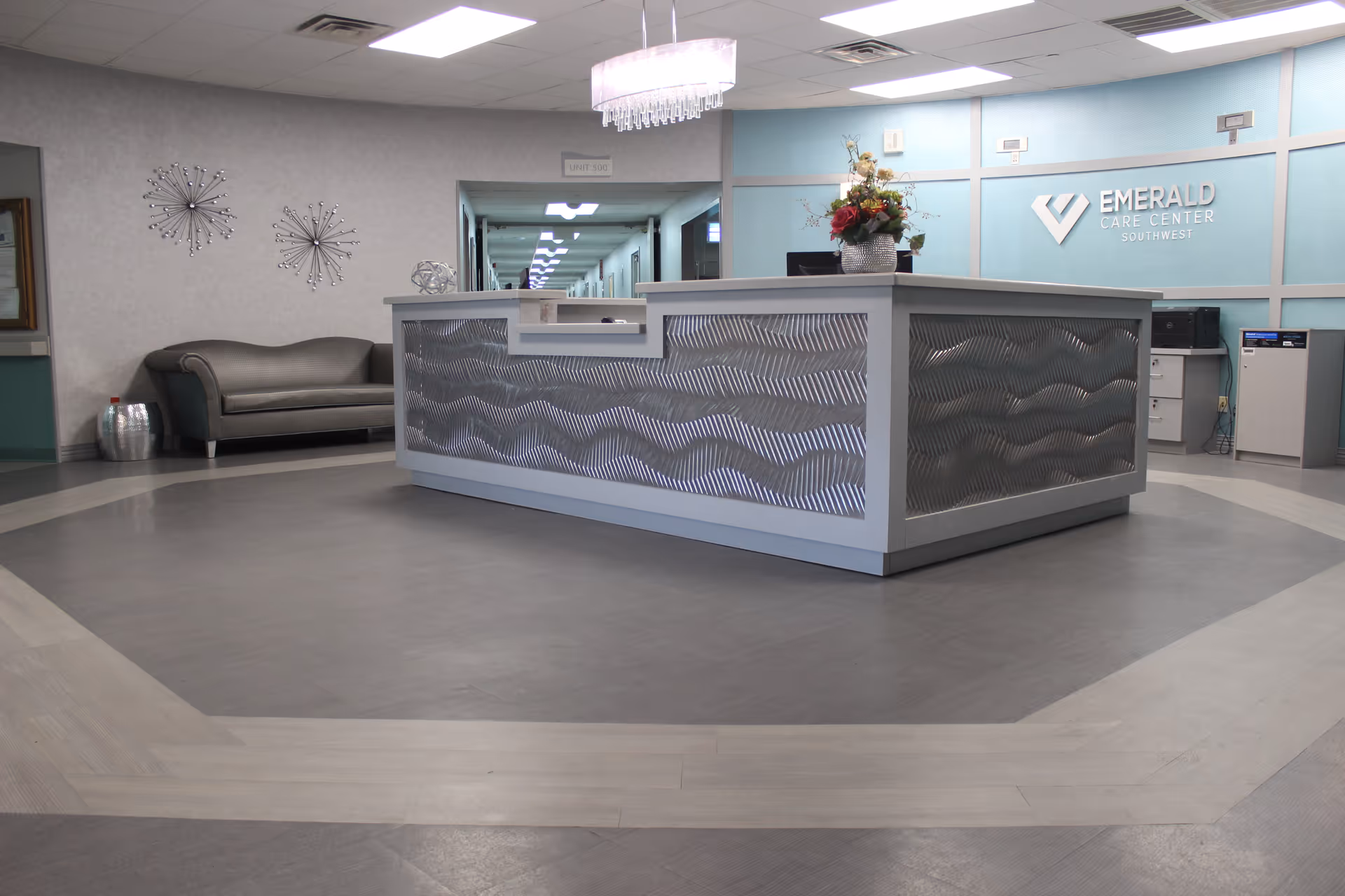 Reception area of Emerald Care Center Southwest featuring a modern front desk with a decorative wave pattern, a flower arrangement on the desk, a gray couch against the wall, two decorative wall hangings, and a hallway leading to other rooms.