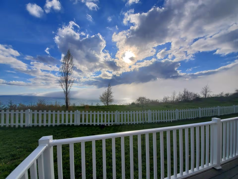 View of a grassy yard enclosed by a white picket fence with a few trees and a large body of water in the background under a partly cloudy sky with the sun shining through.