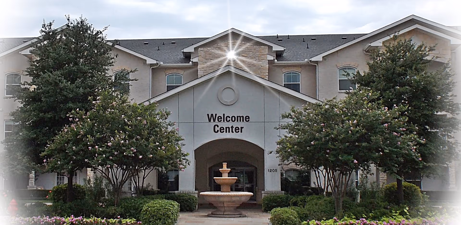 Exterior view of a building labeled Welcome Center with a stone fountain in front, surrounded by trees and bushes under a cloudy sky.