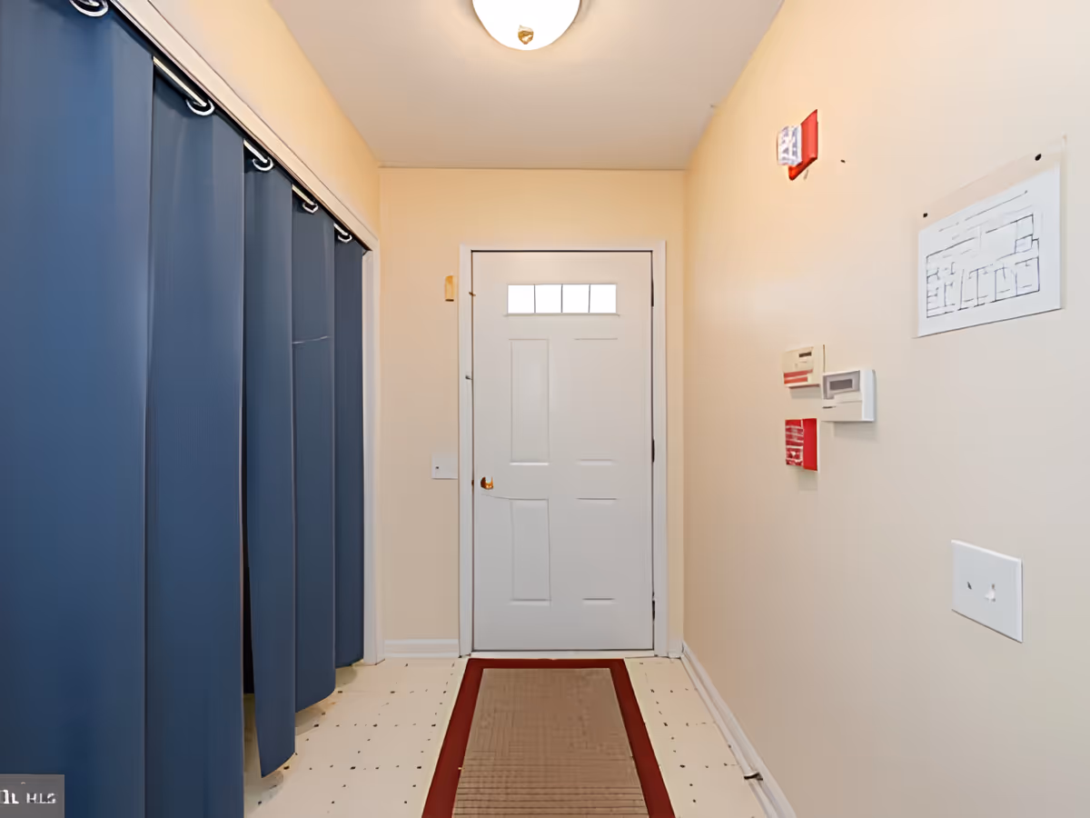Small interior entry hallway with a closed white door, blue curtains on the left, beige walls, and safety devices mounted on the right wall.