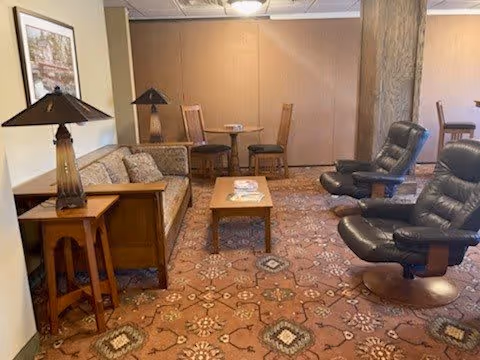 A cozy living room area with a patterned carpet, two black leather recliners, a wooden coffee table with magazines, a beige sofa with cushions, two wooden side tables each with a lamp, and a small round table with two chairs in the background. A framed picture hangs on the wall.