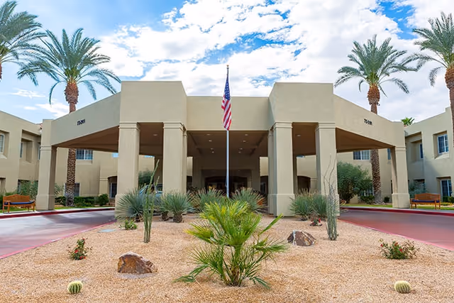 Front exterior view of Bayshire Rancho Mirage facility with a beige building, an entrance canopy supported by columns, an American flag on a flagpole in the center, desert landscaping with palm trees, cacti, and rocks, and a partly cloudy sky.