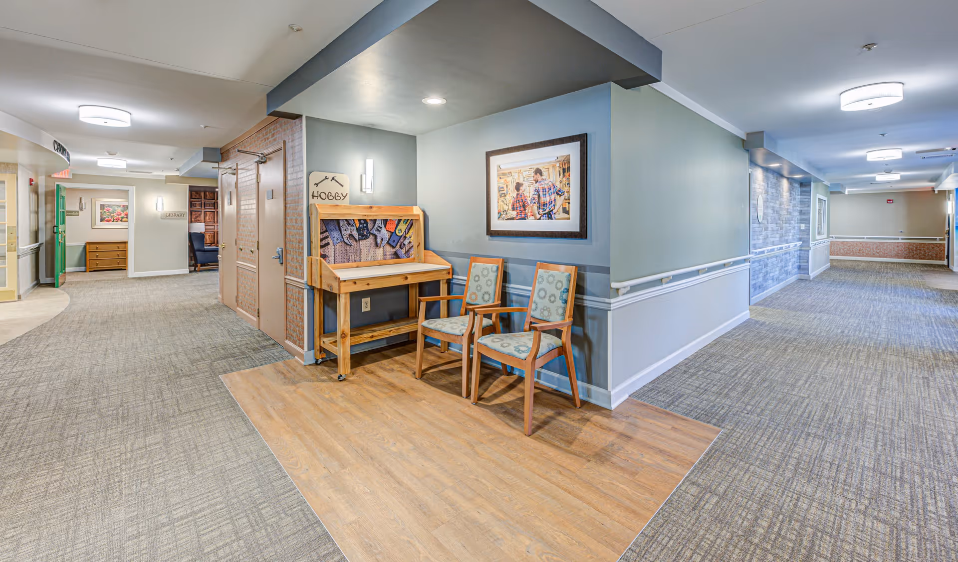 Interior hallway of a senior living facility with carpeted floors and soft lighting. There is a small wooden hobby station with tools and two chairs against a wall, beneath a framed picture of two people. The hallway extends in two directions with doors and signage visible, including a sign for a library.