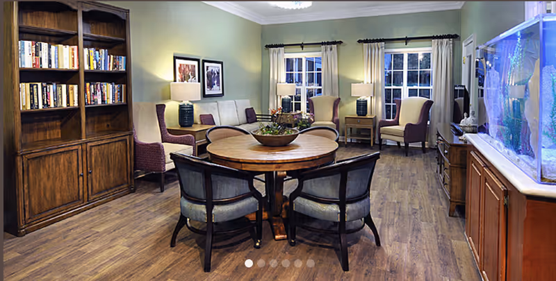 A cozy senior living common area with wooden flooring, a round wooden table surrounded by four cushioned chairs, two armchairs with purple upholstery, a beige sofa, a wooden bookshelf filled with books, two table lamps on side tables, framed artwork on the wall, and a large aquarium on a wooden cabinet.