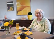 An elderly woman with white hair and glasses sits at a dining table set with plates of food, a glass of iced tea, and a small vase with a yellow flower. Behind her is a sideboard with decorative items and a large yellow flower painting on the wall.