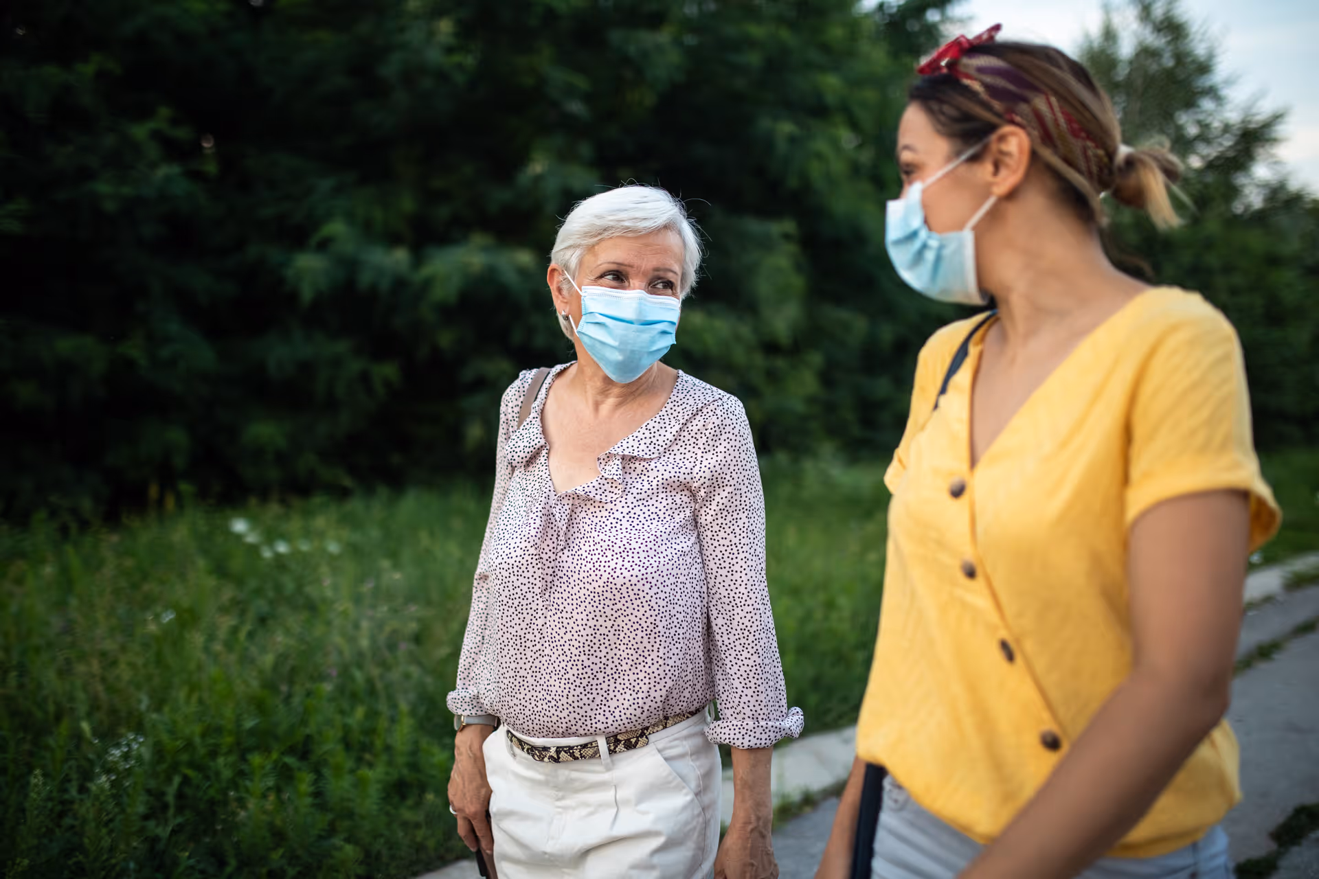 Two women wearing face masks walking outdoors on a path with greenery and trees in the background, engaging in conversation.