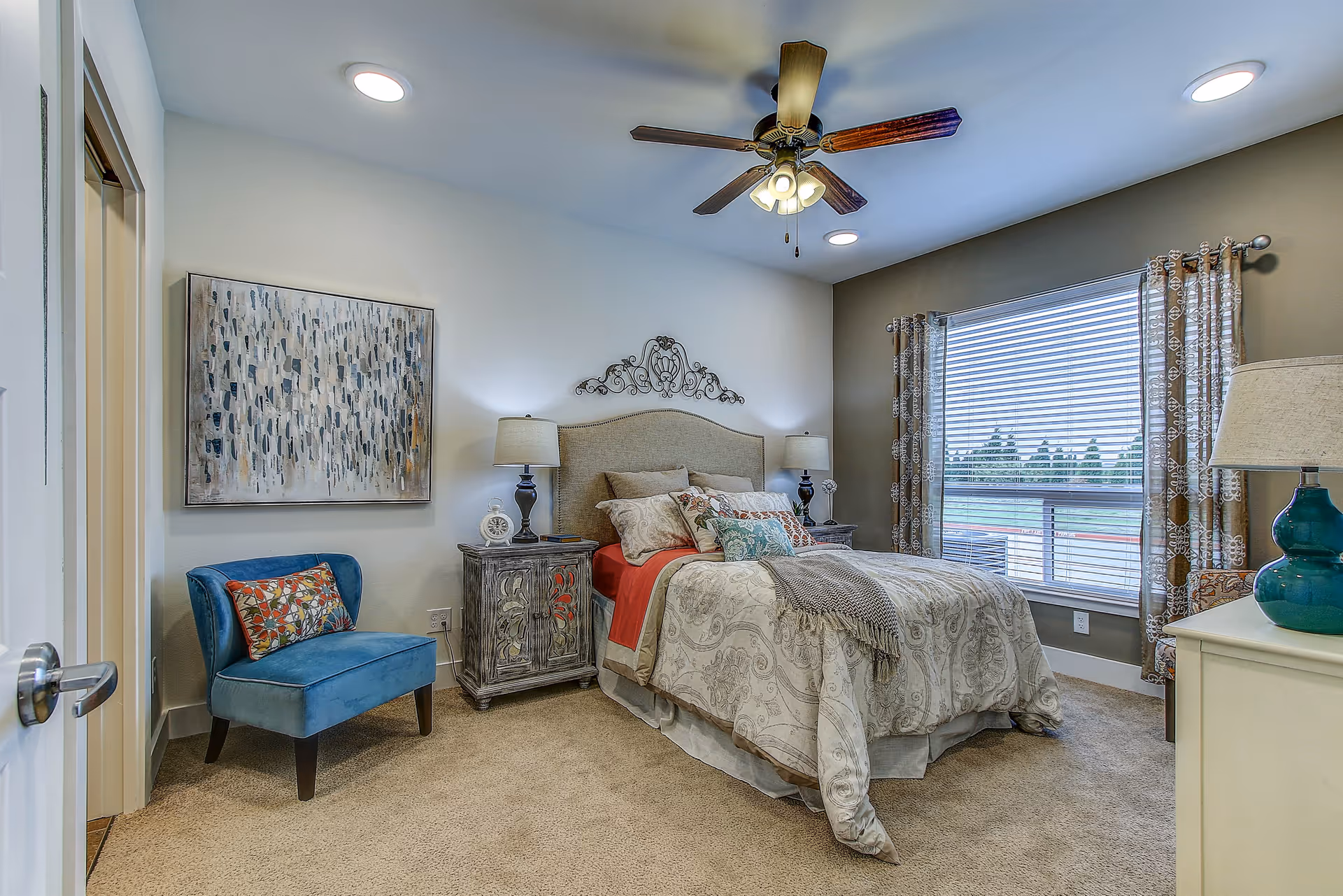 A cozy bedroom with a large bed covered in patterned bedding and multiple pillows. There are two nightstands with lamps on either side of the bed. A blue upholstered chair with a colorful pillow is placed near the door. The room has a ceiling fan with lights, a large window with patterned curtains, and a piece of abstract artwork on the wall.