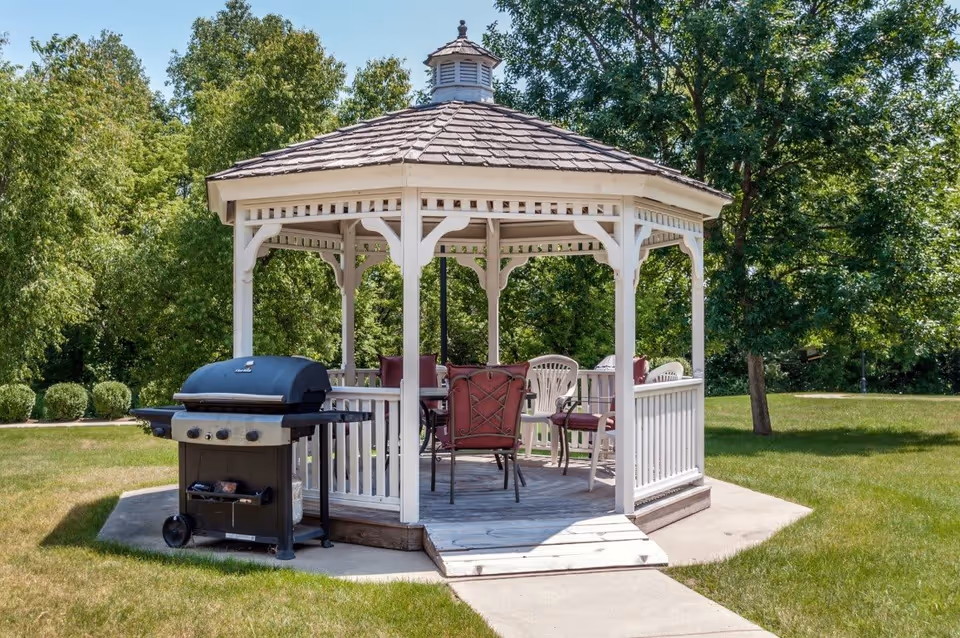 A white wooden gazebo with a shingled roof situated on a concrete pad in a grassy outdoor area. Inside the gazebo are several chairs with red cushions and a table. A black barbecue grill is positioned just outside the gazebo. Trees and bushes surround the area under a clear blue sky.
