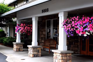 Front entrance of a senior living building with stone columns, a wooden bench, and hanging pink and purple flower baskets beneath the address '9803'.