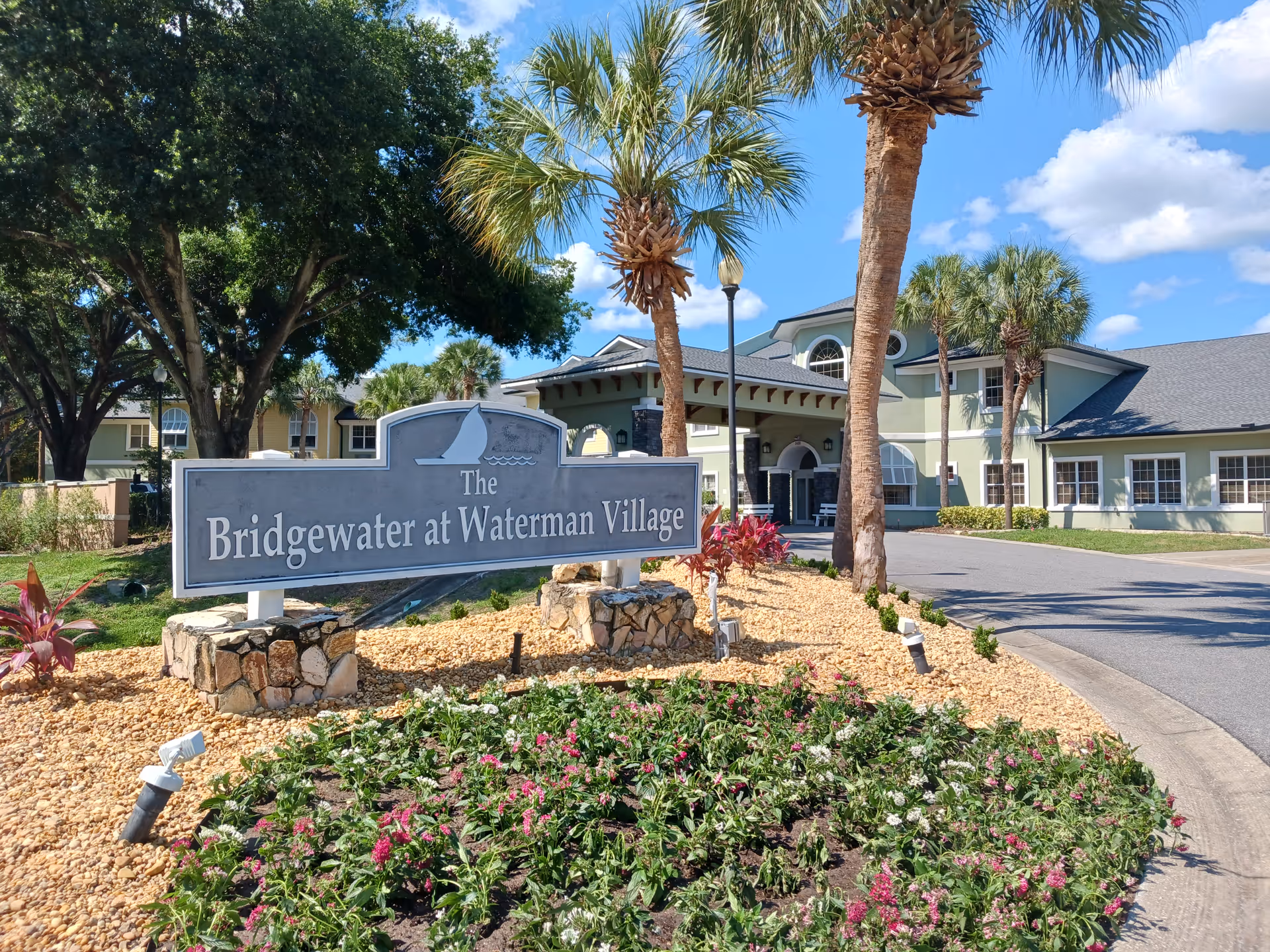 Entrance sign for Bridgewater at Waterman Village with landscaped flowerbeds, palm trees, and the facility building in the background under a blue sky.