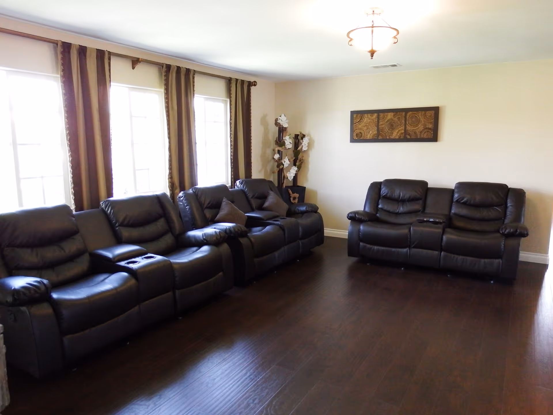 A living room with two black leather reclining sofas arranged against cream-colored walls. There are three large windows with brown and beige curtains on the left side, and a decorative wall art piece above one sofa. A tall plant with white flowers is placed in the corner near the wall art. The floor is dark wood, and a ceiling light fixture is visible.