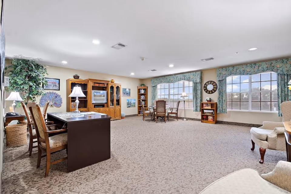 A spacious living room area with carpeted flooring, multiple chairs, a wooden entertainment center with a TV, large windows with floral curtains, a clock on the wall, and several lamps providing warm lighting.