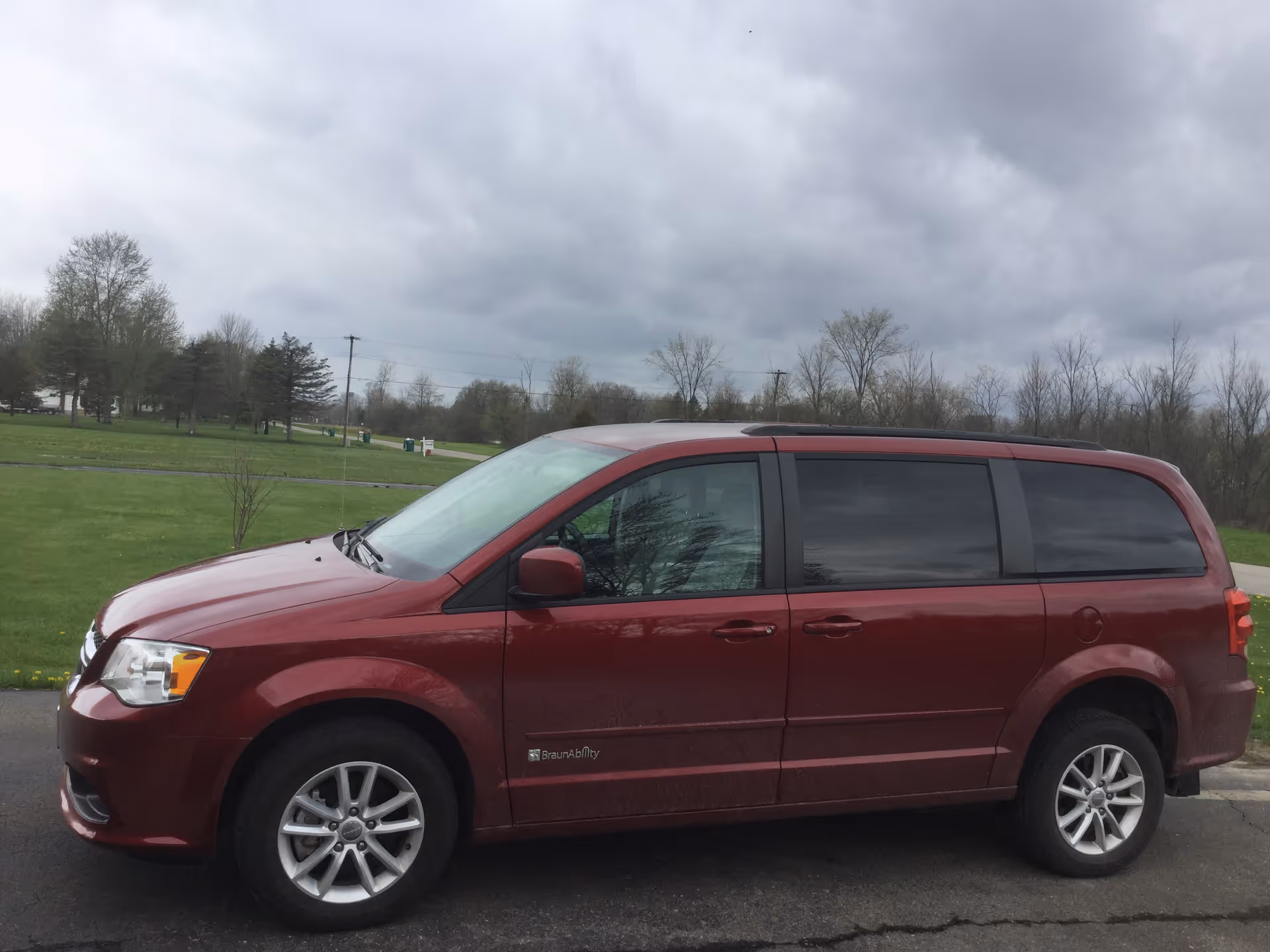 A maroon minivan parked on a paved surface with a grassy area and trees in the background under a cloudy sky.