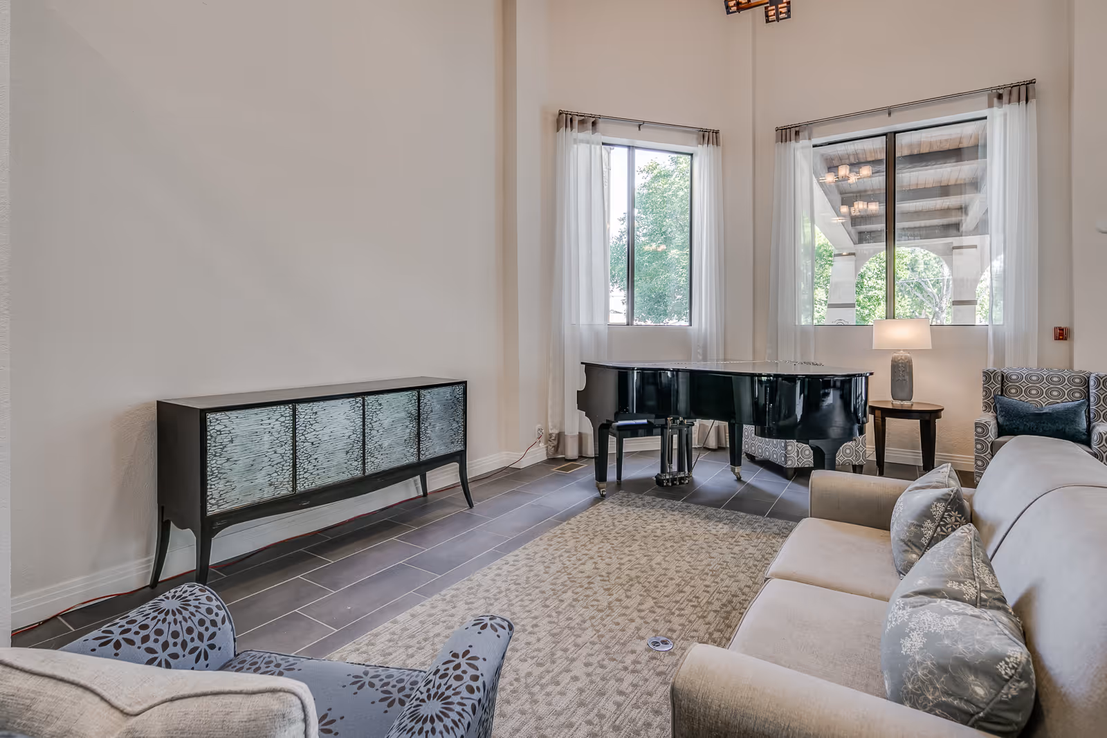 A bright living room with a beige sofa adorned with patterned pillows, a patterned armchair, a black grand piano near two large windows with sheer white curtains, a side table with a lamp, and a decorative cabinet against the wall. The room has tiled flooring and a light-colored area rug.