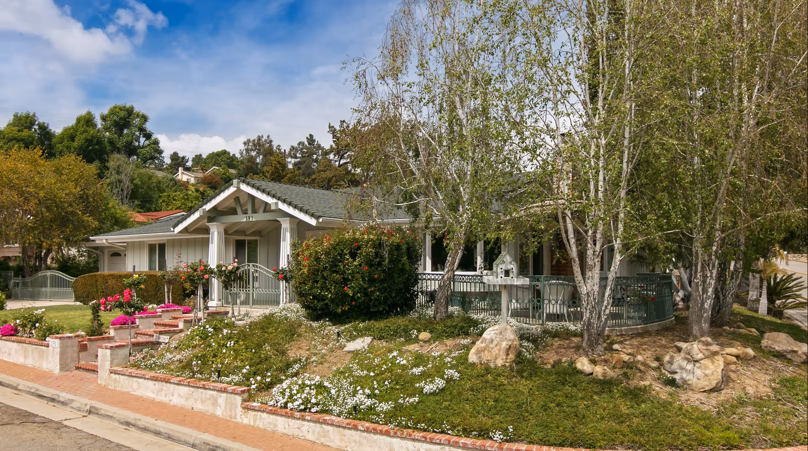 Exterior view of a single-story senior care facility building with a front porch, surrounded by trees, bushes, and flowering plants under a partly cloudy sky.