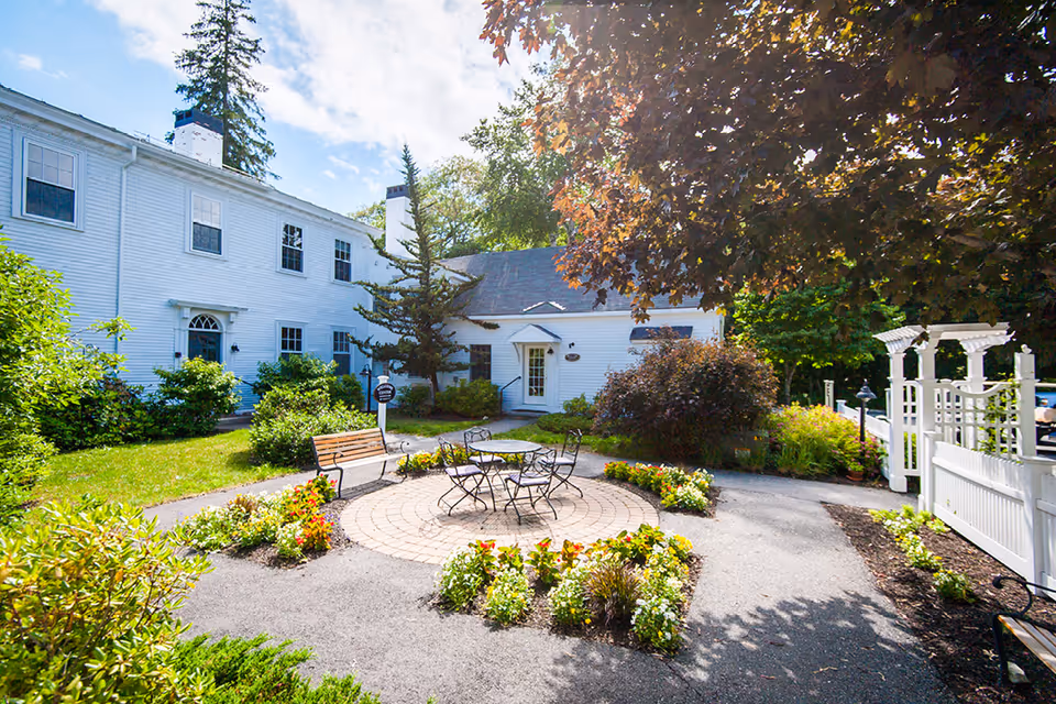 Outdoor courtyard area at The Highlands featuring a circular paved seating area with a metal table and chairs, surrounded by flower beds and greenery. There are benches along the pathway, white fencing, and a white two-story building with multiple windows in the background under a partly cloudy sky.