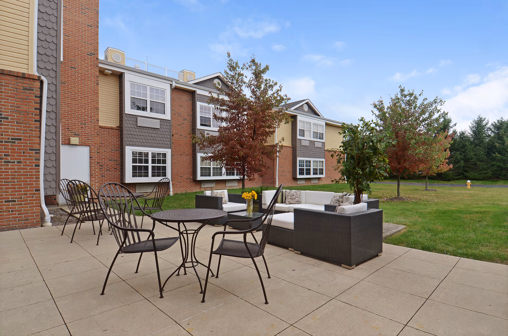 Outdoor patio area at Stoney Ridge Senior Living with metal chairs and tables, a cushioned sectional sofa with pillows, and a small table with a vase of yellow flowers. The patio is adjacent to a brick and siding building with multiple windows, surrounded by grass and trees under a partly cloudy sky.