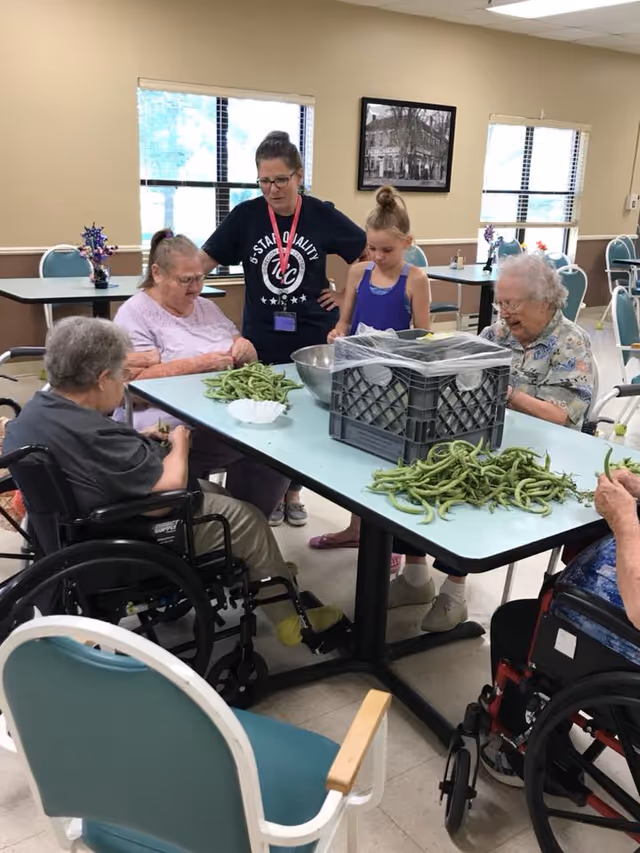 A group of elderly women and a young girl sitting and standing around a table in a communal room, shelling green beans. One woman is in a wheelchair, and a caregiver stands nearby watching. The room has beige walls, windows with blinds, and several chairs and tables.